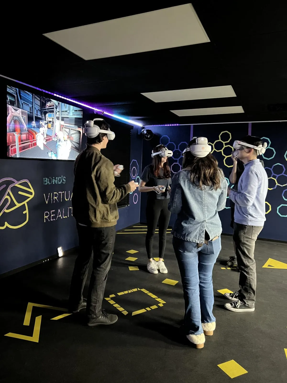 Four adults, two of them in cowboy hats, wearing VR headsets and holding controllers in Bond's free-roam VR arena, under a Bond's Virtual Reality mural