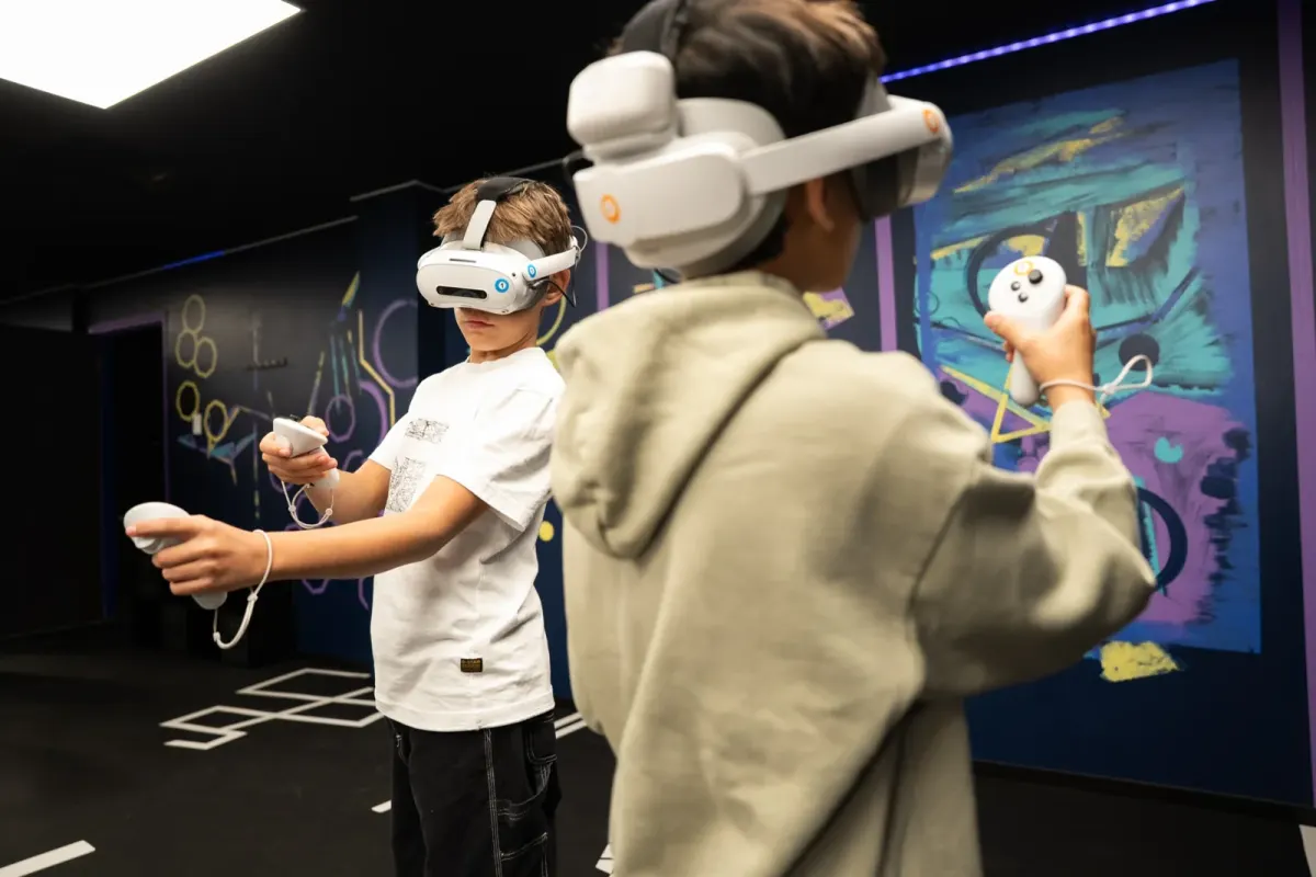 Two boys in white VR headsets holding controllers and playing side by side in front of a painted neon wall