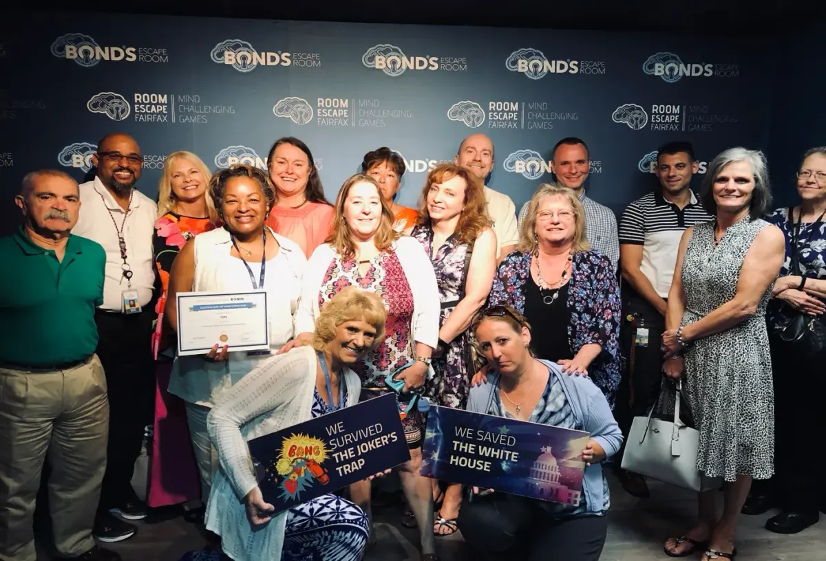 A group of colleagues smiling in front of a Bond's Escape Room Fairfax backdrop, two of them holding signs that read We survived the Joker's Trap and We saved the White House, with a certificate of participation