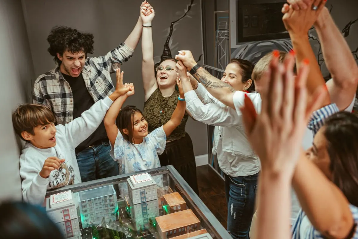 A family with two young children and several adults throw their arms up and cheer over a tabletop model of a white building