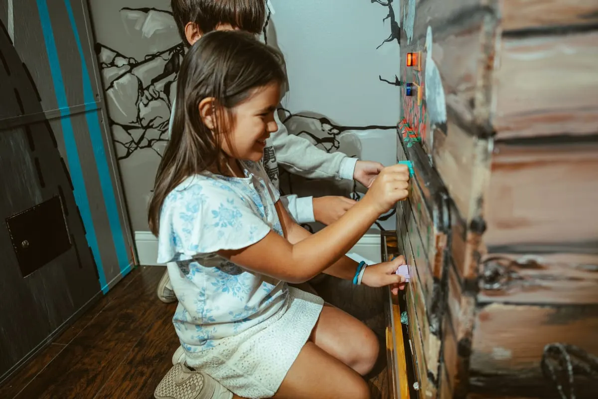 A young girl in a white dress concentrates on a puzzle mounted on a painted wooden wall inside an escape room