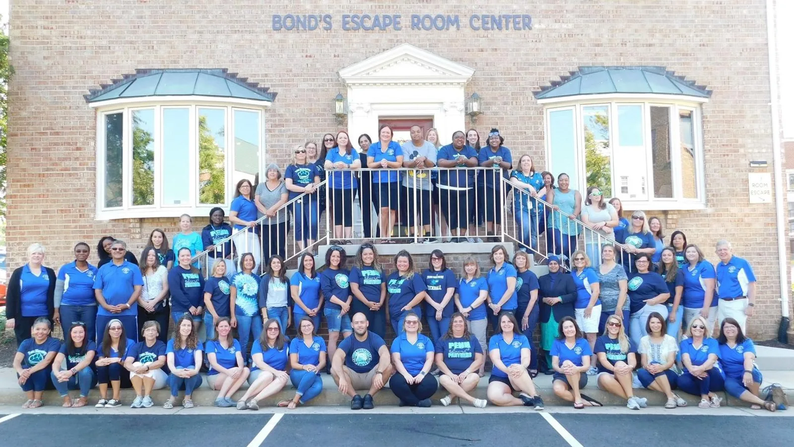 The Bond's team in matching blue T-shirts lined up on the front steps and lawn of the brick Bond's Escape Room Center in Old Town Fairfax