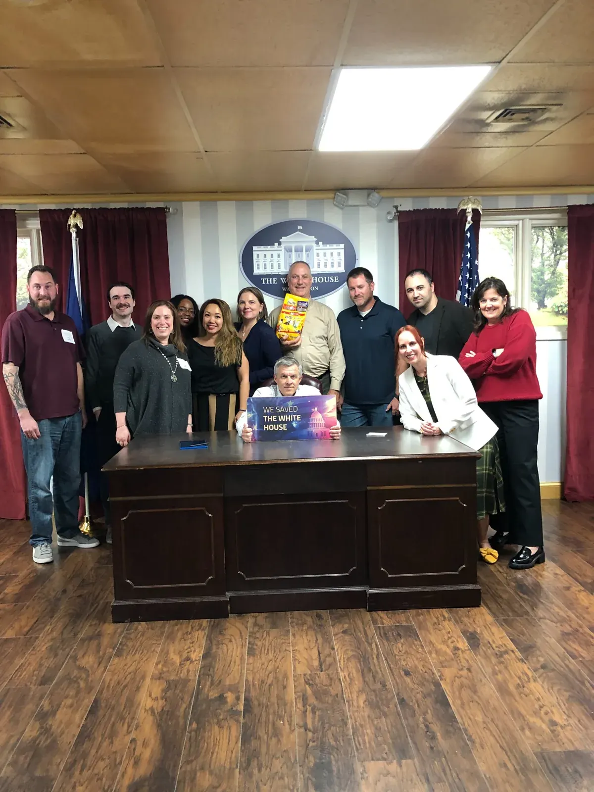 A team of about a dozen colleagues posing behind a desk in the White House escape room set, holding a sign that reads We Saved the White House