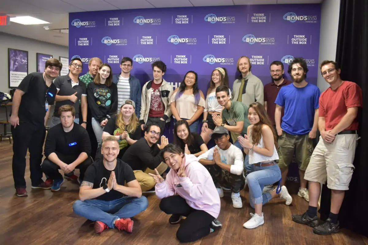About twenty people in casual clothes posing close together, some kneeling in front, with a purple Bond's Escape Room Think Outside the Box banner behind them