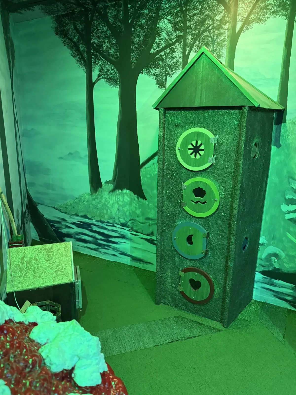 A carved wooden tower with round dials and lock panels standing among painted trees, lit green