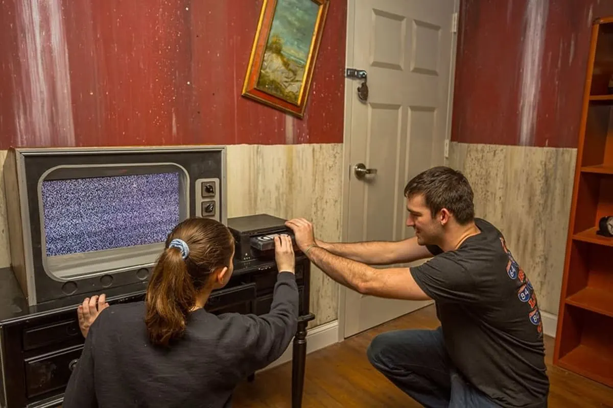 Two players crouched at an old television showing static, working at the dials together in a wood-panelled room