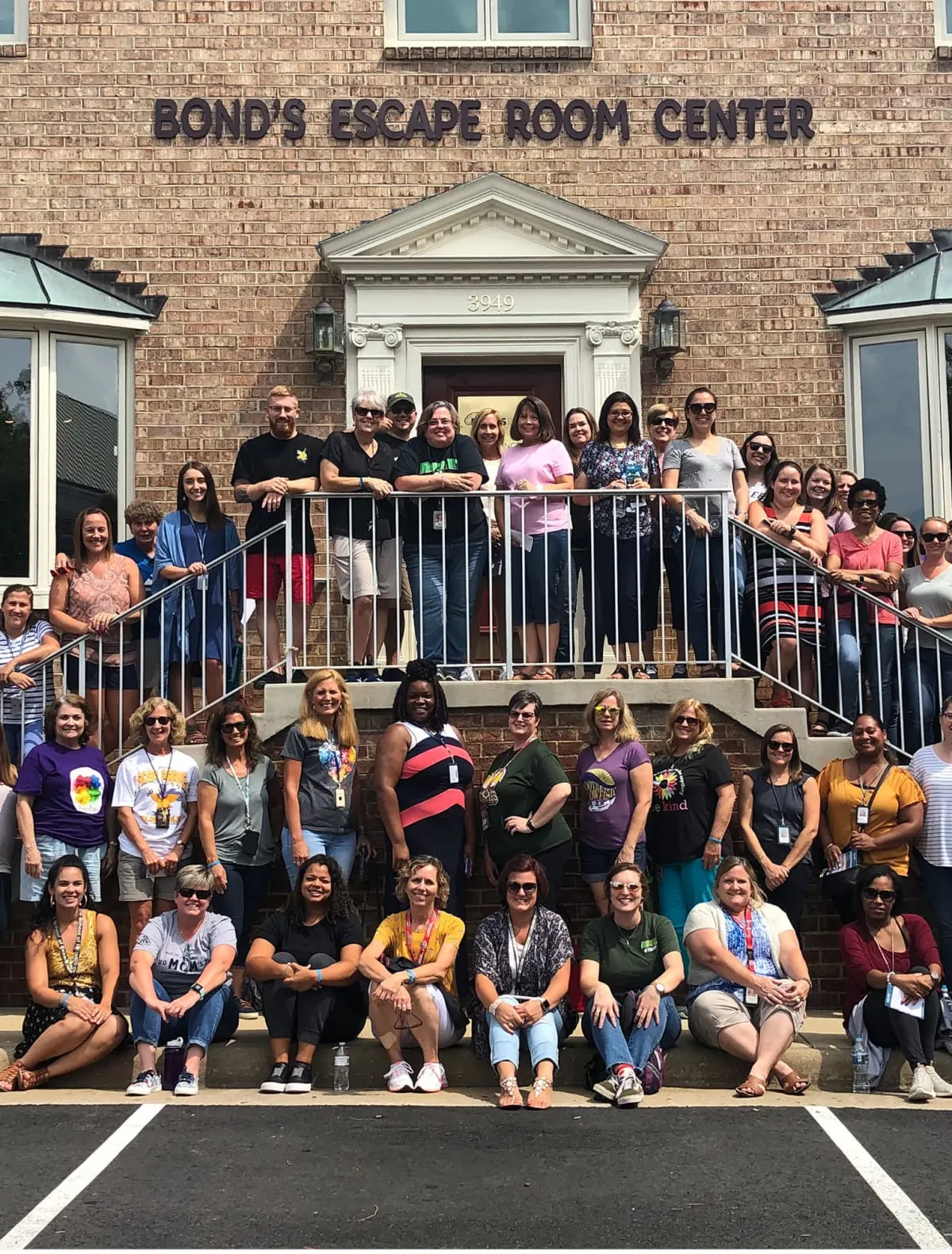 A large company group posing on the front steps of the brick Bond's Escape Room Center building in Fairfax