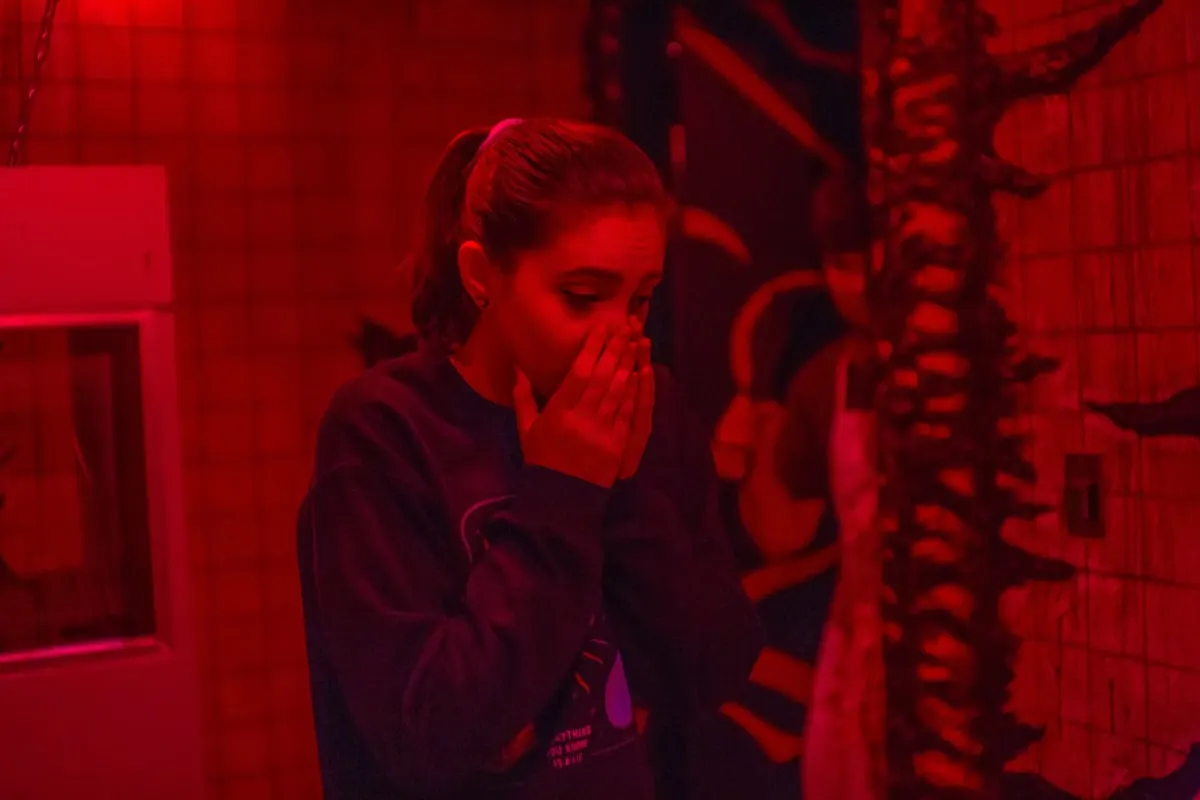 A teenage girl in deep red light with a hand pressed to her mouth in fright, beside a twisted, bone-like column