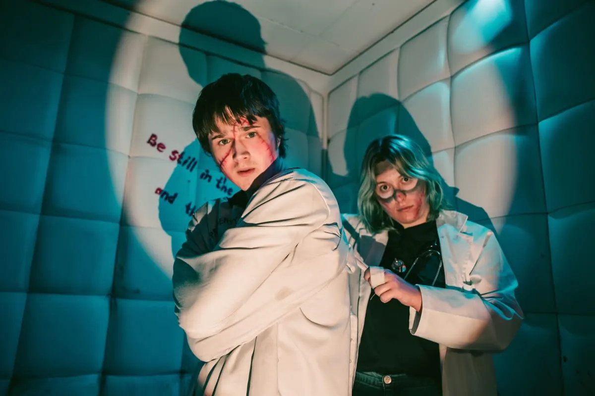 Two players in white lab coats inside a padded cell, lit by a single spotlight, one peering nervously over the other's shoulder