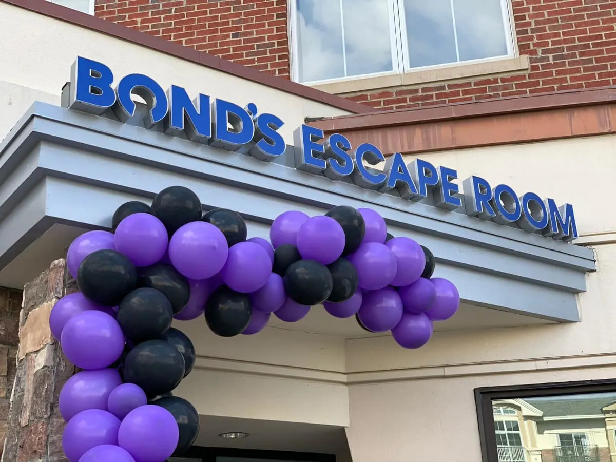 The Bond's Escape Room sign above a building entrance, framed by an arch of purple and black balloons