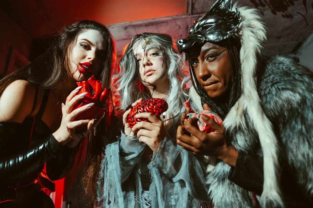 Three people in monster costumes, a vampire, a ghostly bride and a wolf-hooded figure, holding prop hearts and brains in a red-lit room