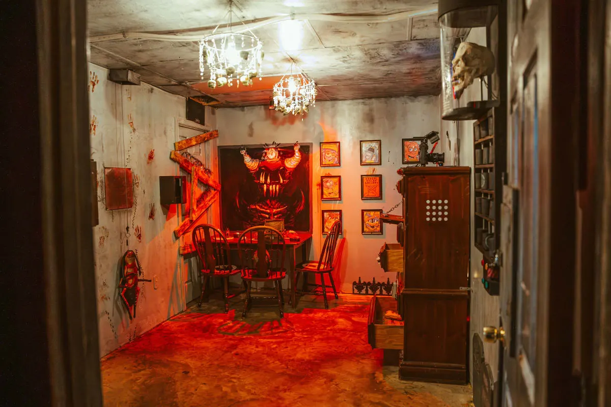 An empty dining room set in red light, with a chandelier, a long table, a skull on a shelf and a demon face painted on the far wall