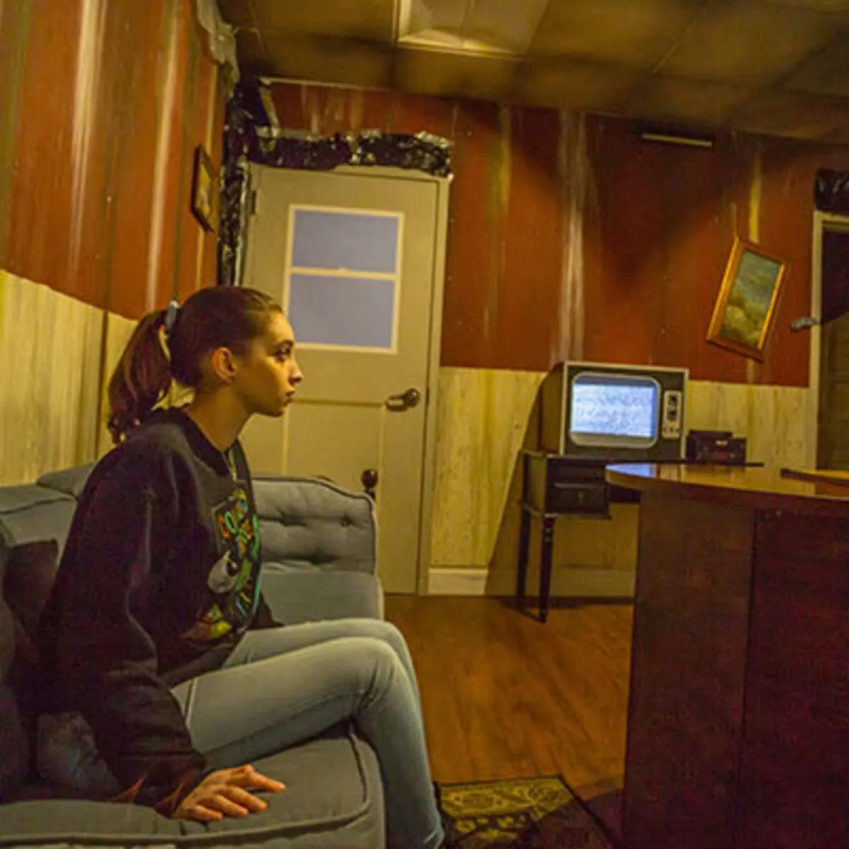A teenage girl sitting on a couch in a wood-panelled den with an old television set and a closed door behind her