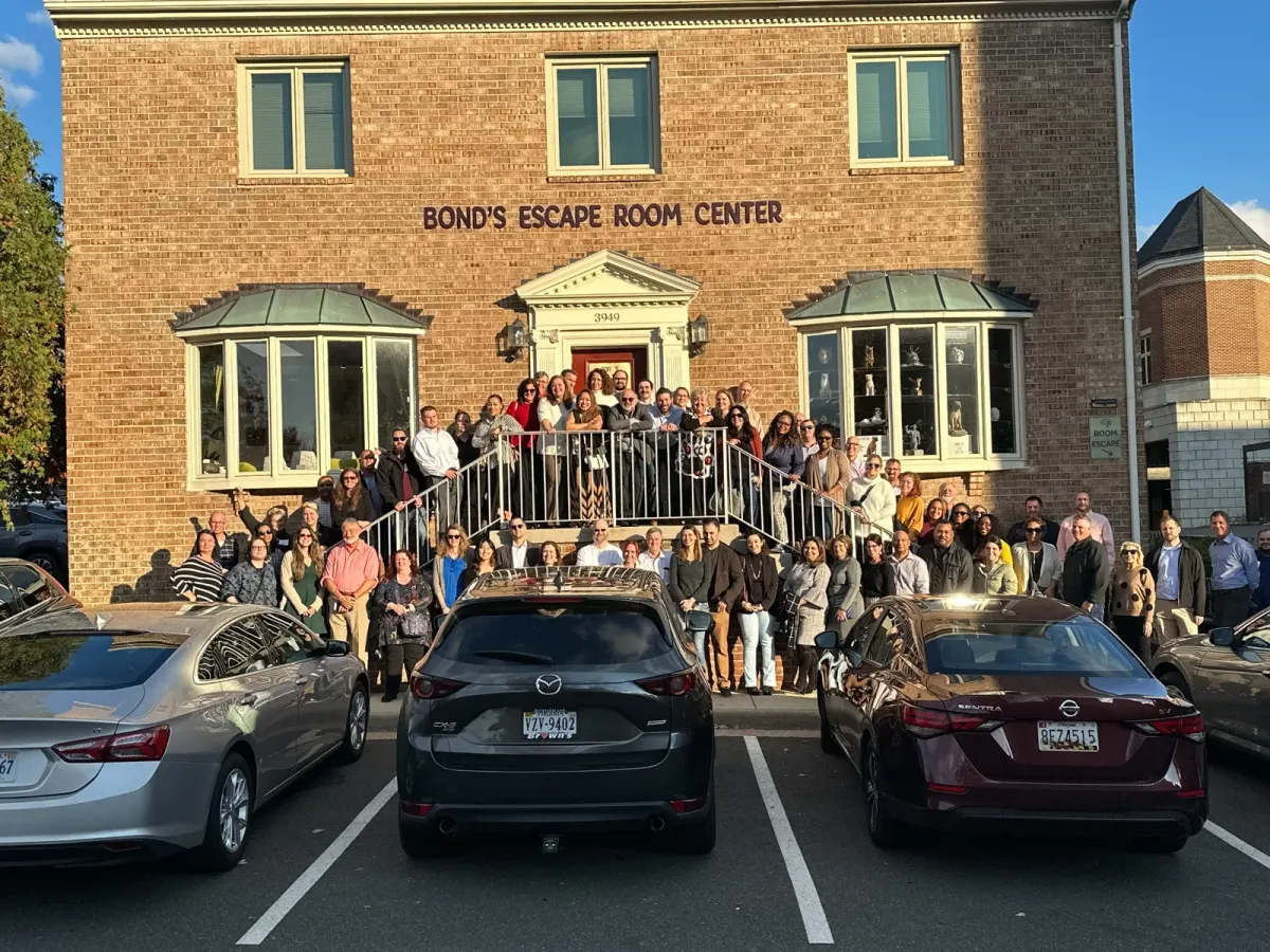 A large group of several dozen people posing on the front steps and the parking lot below the brick Bond's Escape Room Center building in Fairfax