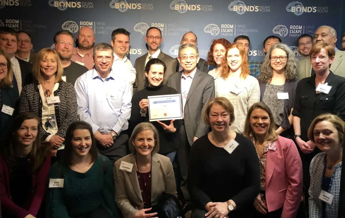 About two dozen colleagues of mixed ages in business and casual dress posing in rows, one holding a framed certificate, in front of a Bond's Escape Room backdrop
