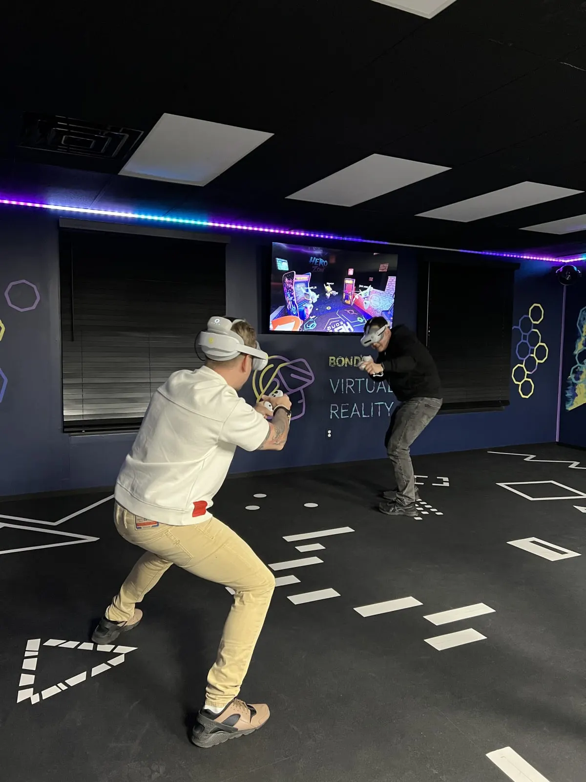 Two players in white VR headsets crouching and aiming controllers inside Bond's free-roam VR arena in Fairfax, with game footage on a wall screen behind them