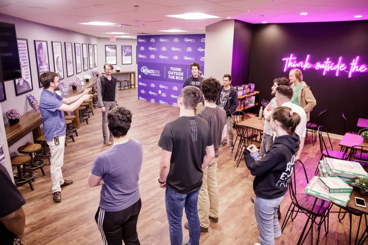 A Bond's host briefing a group of about a dozen people in the lobby, beside a purple Think outside the box backdrop and a pink neon sign, with pizza boxes on a table