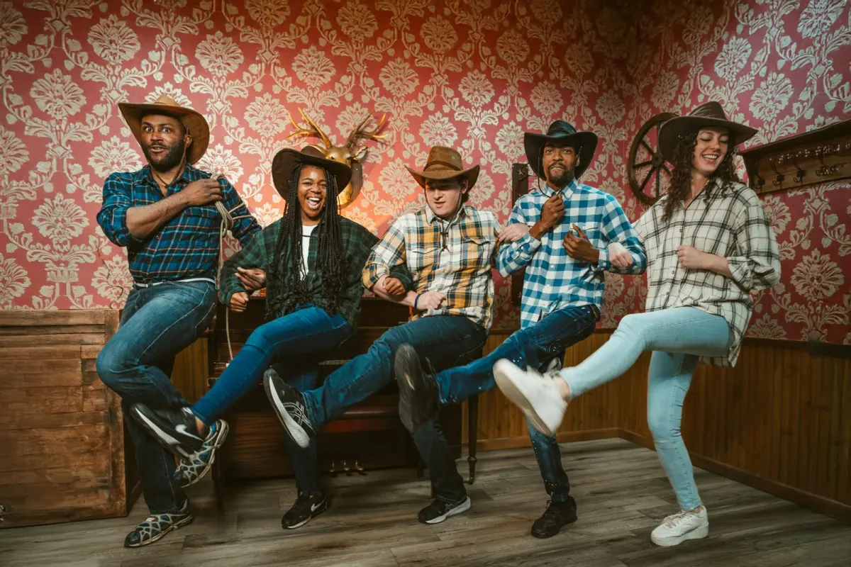 Five coworkers in cowboy hats and plaid shirts linking arms and high-kicking together in a Western-themed room with red damask wallpaper, laughing