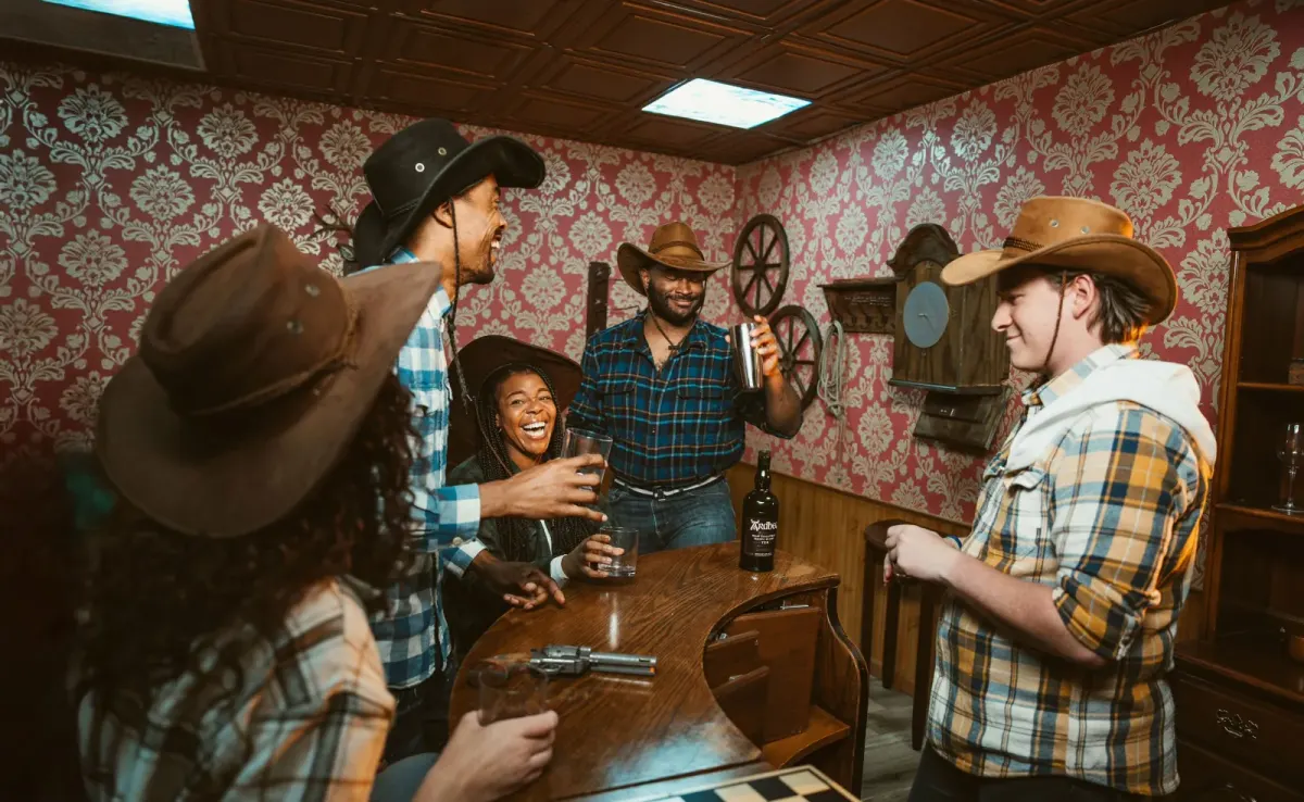 A group in cowboy hats laughing and raising glasses around a curved wooden bar in a Western-themed room with patterned red wallpaper and wagon wheels on the wall