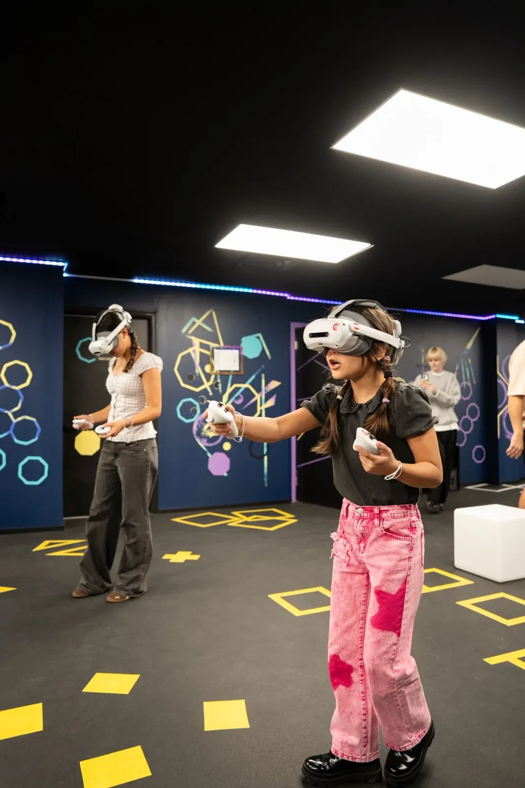 A young girl in a VR headset aiming a controller on a VR arena floor marked with yellow tiles while other players stand behind her