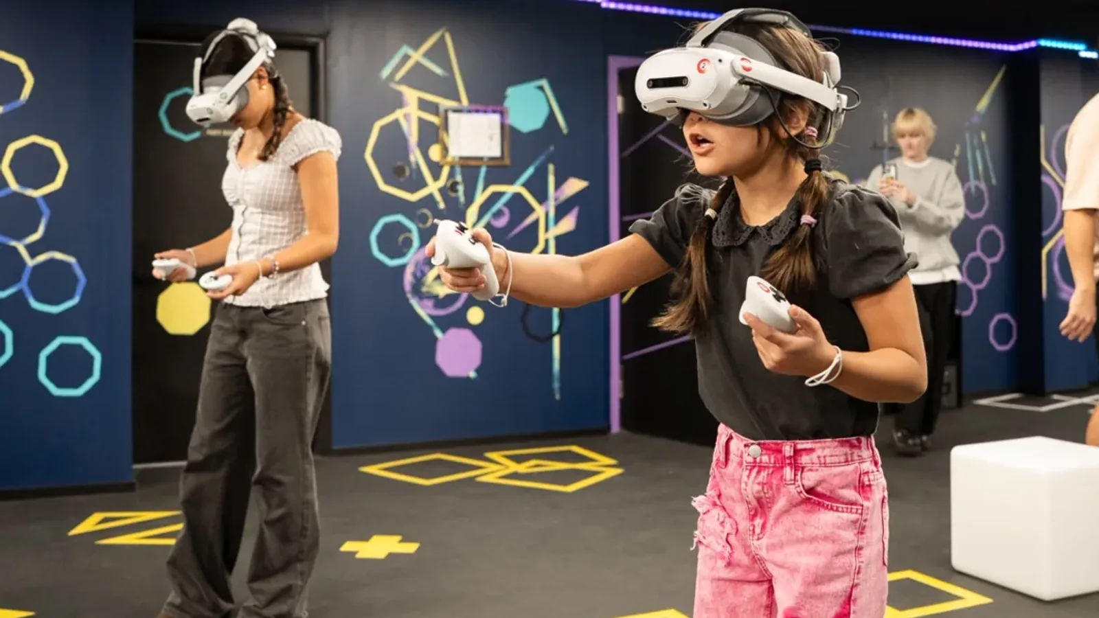 A young girl in a VR headset aiming a controller on a VR arena floor marked with yellow tiles while other players stand behind her