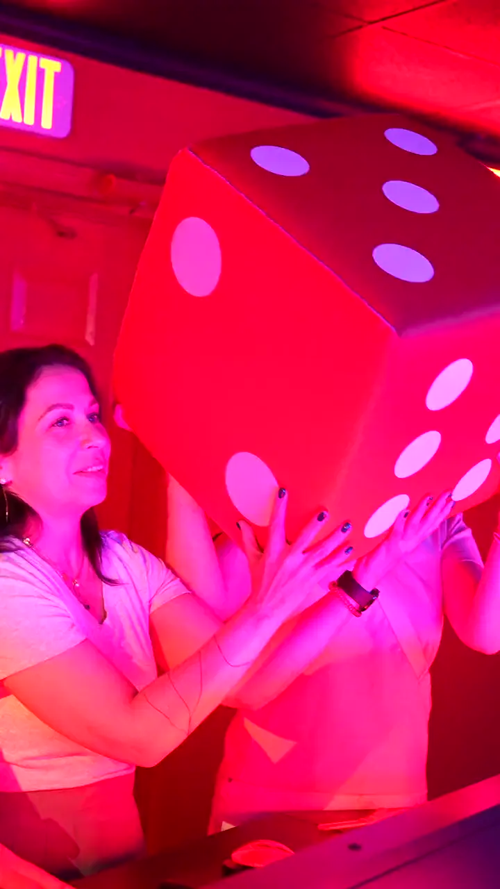 A player under red stage light holding a giant pink die on the Bond's Game Show set