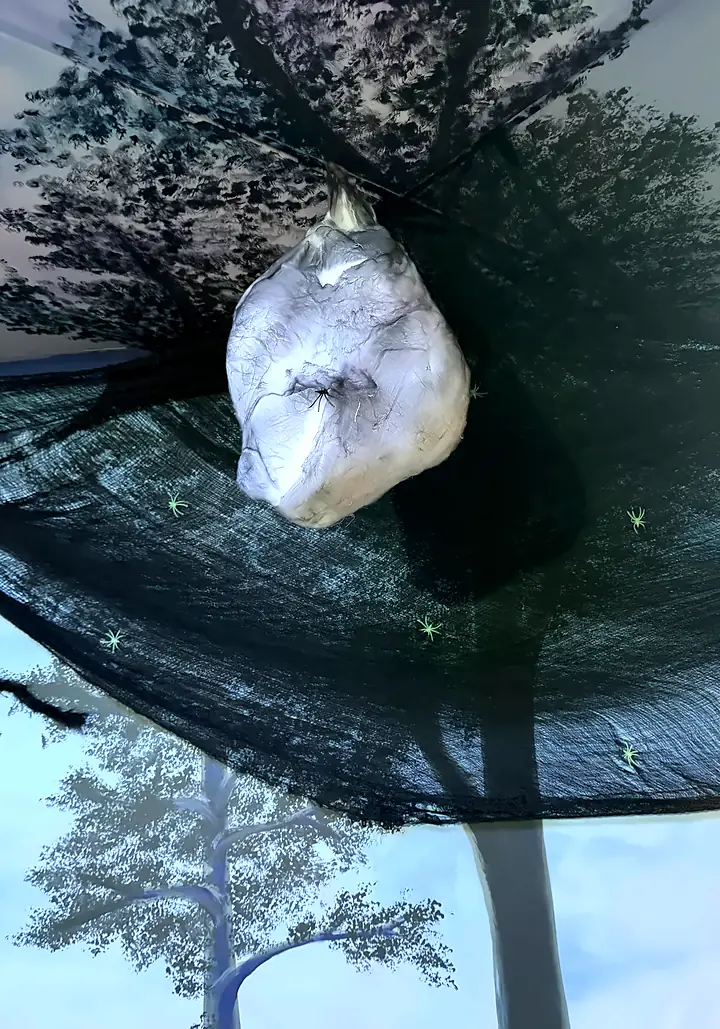 A silver cocoon hangs from a black net canopy in front of the painted forest in The Crawling Dark