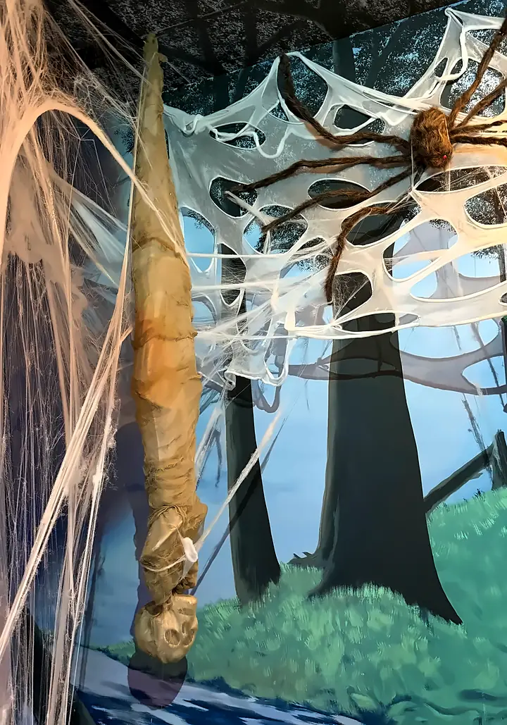 A giant spider on its web beside a hanging cocoon, against the painted forest
