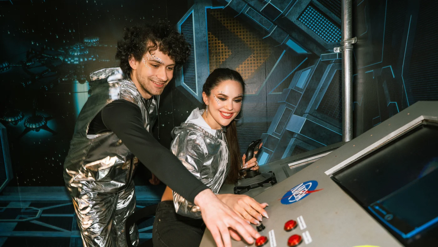 Two players in silver space suits lean over a spaceship console and press red buttons