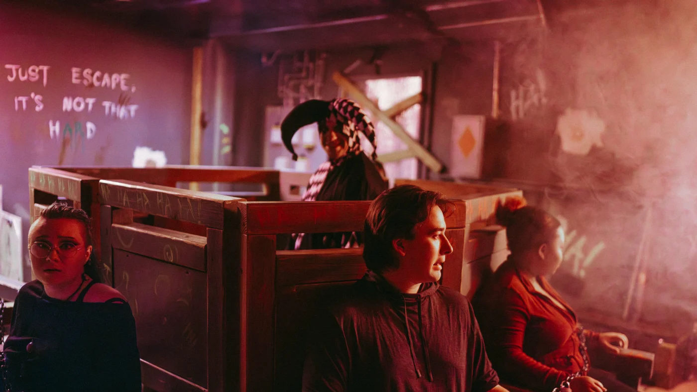 Players sit chained to wooden chairs in a red-lit basement with graffiti on the wall and a clown figure behind them in Survive the Joker's Trap